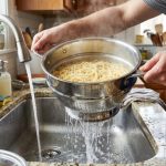 Shiny stainless steel pasta strainer with riveted handles and punched holes draining spaghetti over a kitchen sink