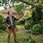 Homeowner using a cordless pole saw to trim high tree branches safely from the ground in a garden setting
